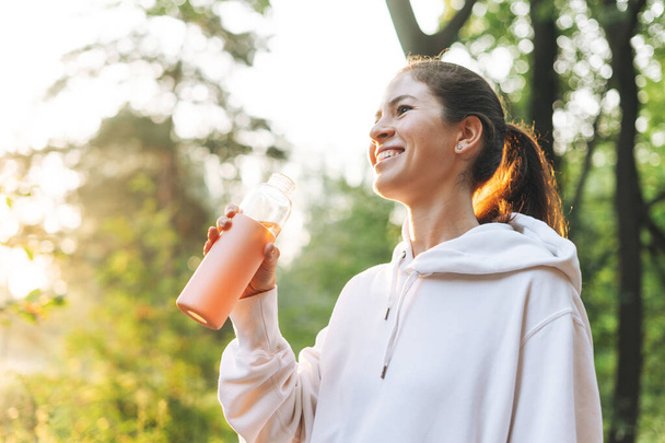 Junge schlanke Frau brünett in Sportkleidung läuft und trinkt Wasser im Wald zur goldenen Stunde Sonnenaufgang. Gesundheit und Wellness, Fitness-Lebensstil - Foto, Bild