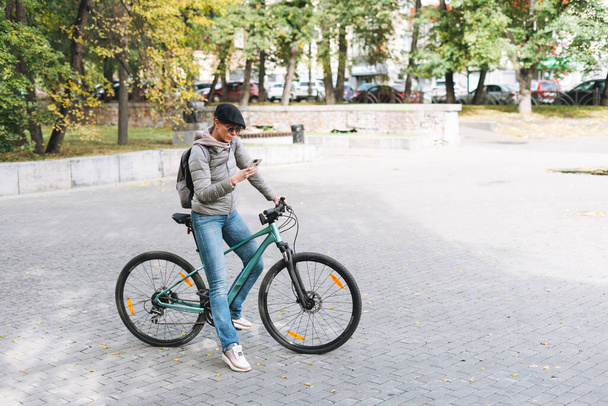 Portrait of fashionable young pretty woman in cap and sunglasses using mobile on bicycle on sunny autumn day in city park - Foto, Imagen