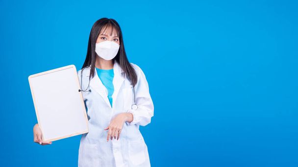 Professione medico o infermiera donna asiatica indossare cappotto medico e maschera protettiva viso con stetoscopio tenendo bordo bianco in mano in piedi sopra isolato sfondo blu. Concetto di protezione sanitaria.  - Foto, immagini