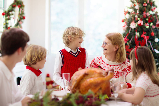 Family with children eating Christmas dinner at fireplace and decorated Xmas tree. Parents and kids enjoy festive meal. Winter holidays celebration and food. Grandmother cooking roasted turkey.  - Photo, Image