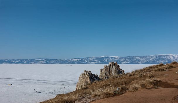 A picturesque two-headed rock, devoid of vegetation, rises above a frozen lake. Cars and tiny silhouettes of people are visible on the ice. In the foreground is a hillside with dry grass. Baikal.  - Photo, Image