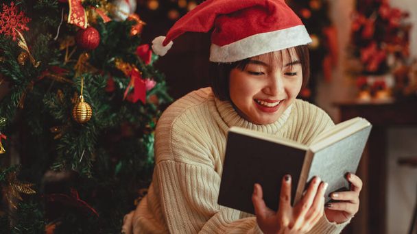 a young Woman in front of Christmas tree reading a book at home with x-mas decoration - Foto, Imagen