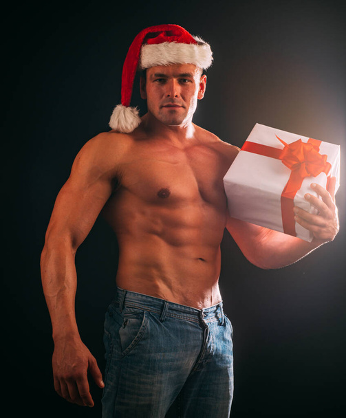 Sexy shirtless Santa with gift on black background. Naked muscular man in Santa hat in studio. Handsome man in Christmas look. Sexy hunk in red Santa hat. - Фото, зображення