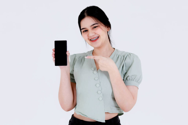 Πορτρέτο στούντιο πλάνο της Ασίας νεαρός έφηβος θηλυκό μοντέλο με αλογοουρά χτένισμα φορώντας οδοντιατρική φροντίδα δόντια τιράντες και των καλλιεργειών κορυφαία στολή στέκεται δείχνοντας κενό smartphone οθόνη σε λευκό φόντο. - Φωτογραφία, εικόνα