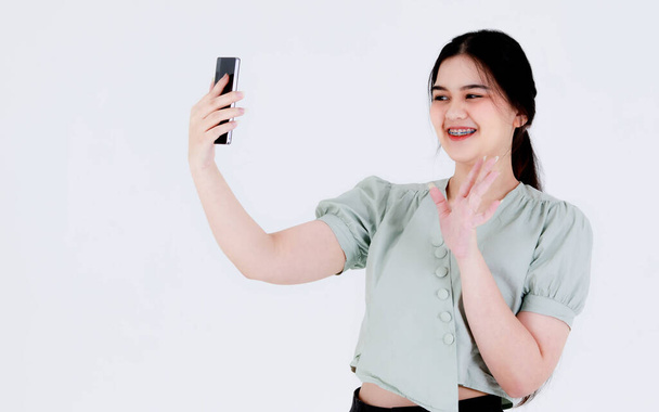Studio shot Azji młody nastolatek kobieta model z kucykiem włosy nosić stomatologiczne opieki zęby aparat ortodontyczny i strój górę zboża machając ręką powitanie powiedzieć cześć na smartphone wideo rozmowy na białym tle. - Zdjęcie, obraz