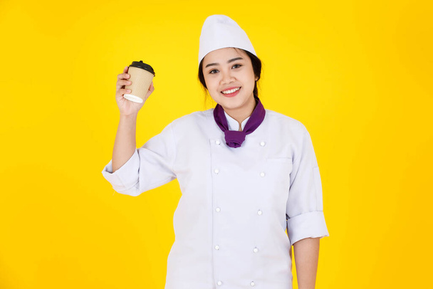 Portrait studio shot of Asian professional successful hotel restaurant female executive chef in white cooking uniform with hat scarf stand smiling holding jetable coffee cup on yellow background. - Photo, image