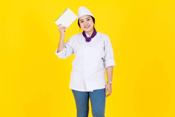 Retrato de estudio de tiro de chef ejecutiva profesional asiática en uniforme de cocina blanco con sombrero y bufanda de pie sonriente celebración de aprender nuevas recetas de cuaderno en las manos sobre fondo amarillo. - Foto, Imagen