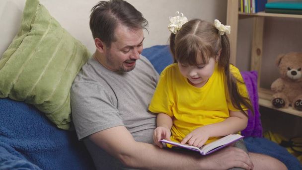 dziecko czyta ciekawą książkę ze swoim ojcem, szczęśliwą rodziną, córeczką odrabiającą lekcje w domu, wesoły dzieciak śmieje się podczas lekcji szkolnych, uczeń szkoły podstawowej uczy się uczyć - Zdjęcie, obraz