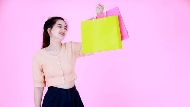Portrait studio shot Asian young happy female shopaholic shopper wearing teeth braces and crop top outfit standing showing presenting colorful shopping bags smiling  on pink background. - Zdjęcie, obraz