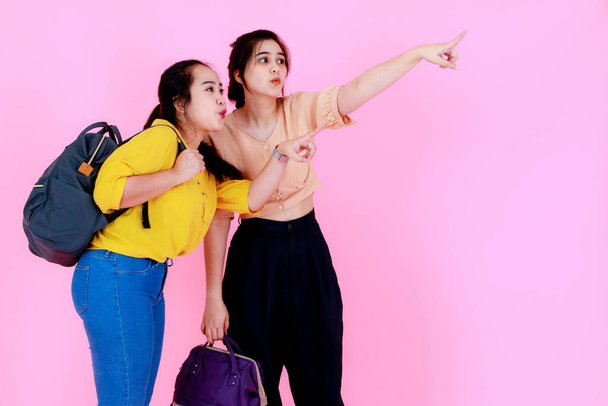 Studio shot van twee Aziatische mollig en slank paardenstaart haar vrouwelijke backpacker vrienden of zussen model in casual slijtage opknoping rugzak op schouder reizen in de natuur wijzend weg samen op roze achtergrond. - Foto, afbeelding