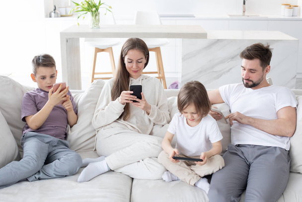 Caucasian parents and kids use devices together sit on sofa, tech addicted family with children hold phone digital tablet having fun with gadgets at home, technology dependence - Zdjęcie, obraz