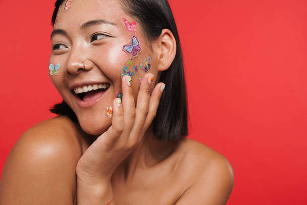 Beauty portrait of a smiling happy young brunette asian woman with makeup facial decoration posing over red wall background - Φωτογραφία, εικόνα