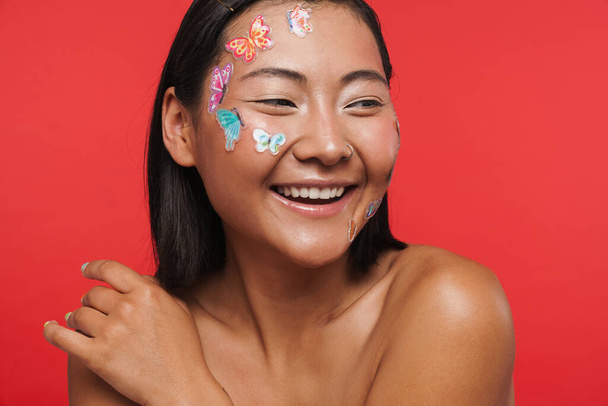 Beauty portrait of a smiling happy young brunette asian woman with makeup facial decoration posing over red wall background - Photo, Image