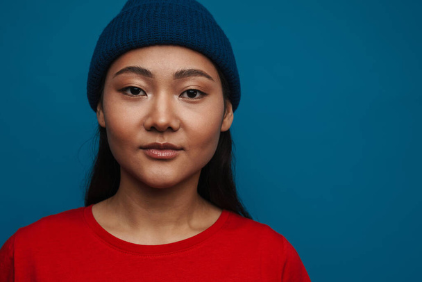 Portrait of a casual young asian woman wearing hat standing over blue wall background - Foto, imagen