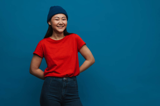 Portrait of a happy casual young asian woman wearing hat standing over blue wall background looking at camera - Foto, afbeelding