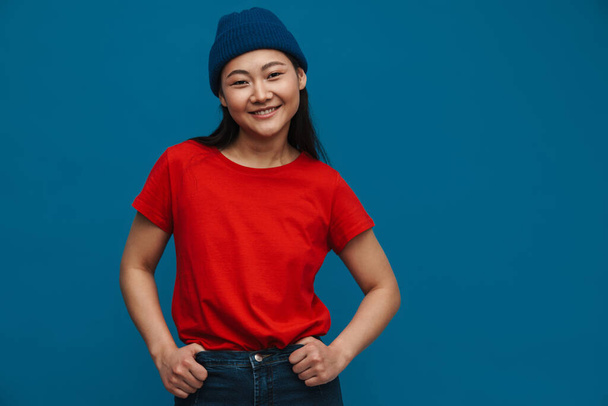 Portrait of a happy casual young asian woman wearing hat standing over blue wall background looking at camera - Fotoğraf, Görsel
