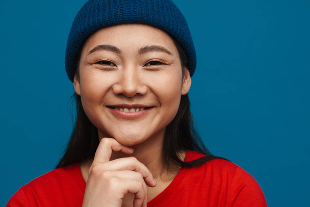 Portrait of a happy casual young asian woman wearing hat standing over blue wall background looking at camera - Foto, imagen