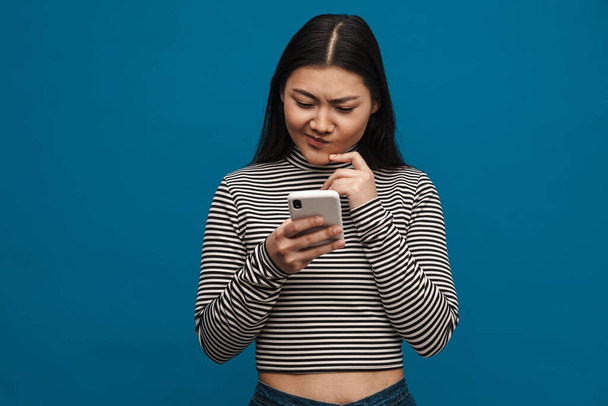 Porträt einer nachdenklichen jungen asiatischen Frau, die vor blauem Wandhintergrund steht und ihr Handy in der Hand hält - Foto, Bild