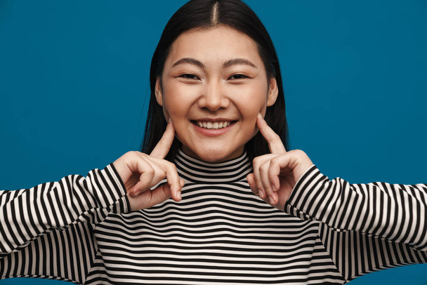 Portrait of a happy casual young asian woman standing over blue wall background looking at camera touching face - Photo, image