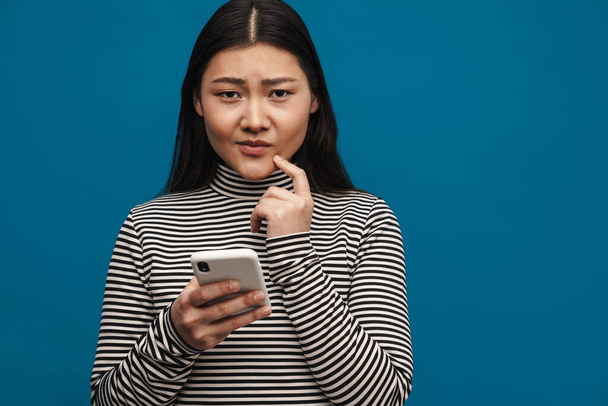 Portrait d'une jeune femme asiatique décontractée sournoise debout sur fond de mur bleu tenant téléphone mobile - Photo, image