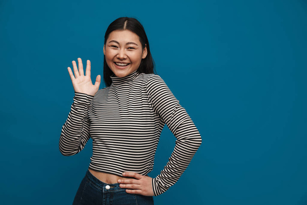 Portrait of a happy casual young asian woman standing over blue wall background waving hand - Foto, Bild
