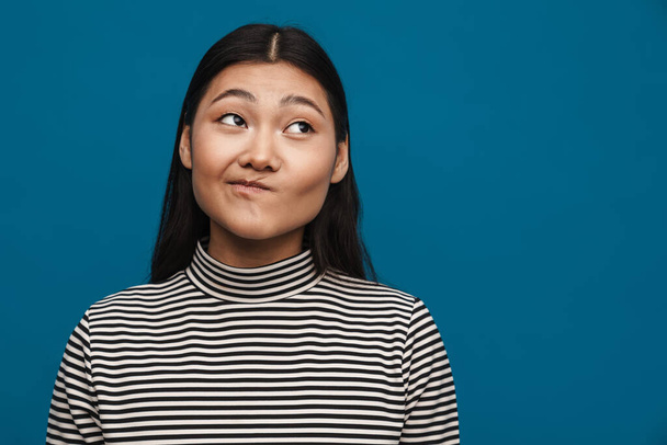 Portrait of a confused casual young asian woman standing over blue wall background looking aside - Valokuva, kuva