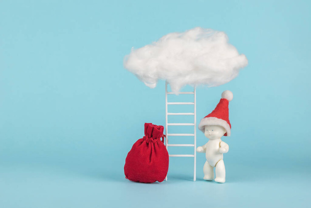 Una figura de un bebé con un sombrero rojo y una escalera que descansa sobre una nube. El concepto de sorpresas de Santa Claus. La tendencia del Año Nuevo. Minimalismo. - Foto, Imagen