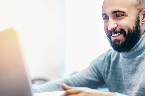 Young man with beard looks at laptop screen and smiles. Internet dating or communication through video communication. - Photo, Image