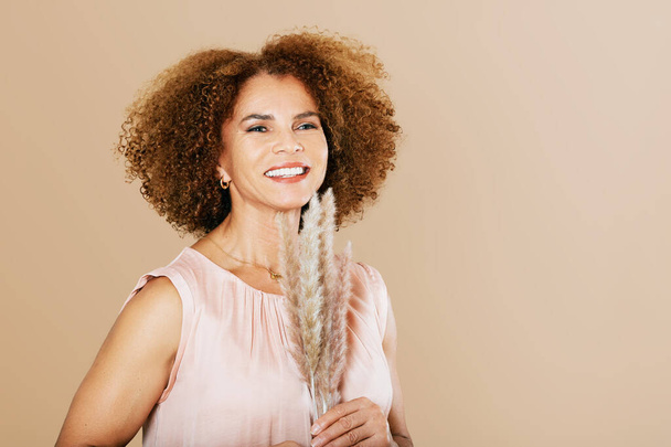 Retrato de estudio de moda de mujer elegante de mediana edad posando sobre fondo beige, señora de 50 - 55 años con cabello rizado sosteniendo hierba pampeana al lado de la cara - Foto, imagen