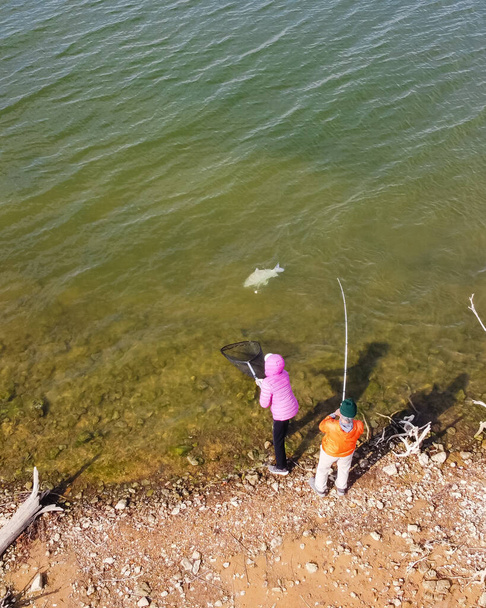 Αεροφωτογραφία ηλικιωμένου ζευγαριού που ψαρεύει και περιμένει να προσγειώσει ένα μικρό βουβάλι από τη λίμνη στο Τέξας των ΗΠΑ. Συνταξιούχοι ενήλικες σε ελαφρά σακάκια με καπέλα beanie απολαμβάνουν δροσερό καιρό υπαίθριες δραστηριότητες - Φωτογραφία, εικόνα