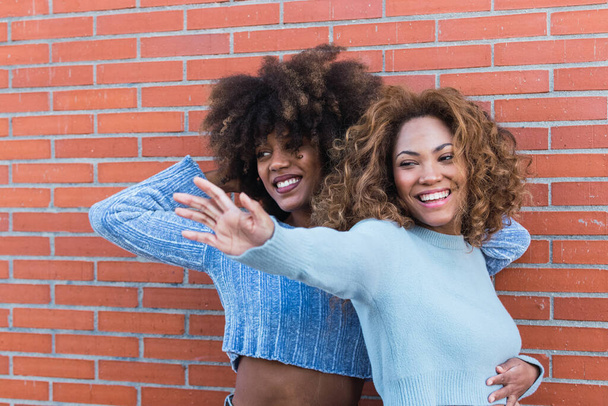 Side view of smiling african american friends with long afro hair, glamour makeup and blue jumper pulling hand towards. Beauty portrait of african natural girls having fun in brick wall - Zdjęcie, obraz