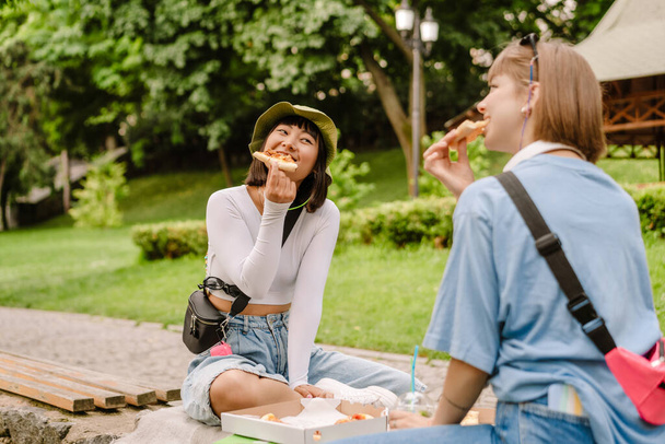 Multiracial two women smiling and eating pizza while resting in green park - Foto, Bild