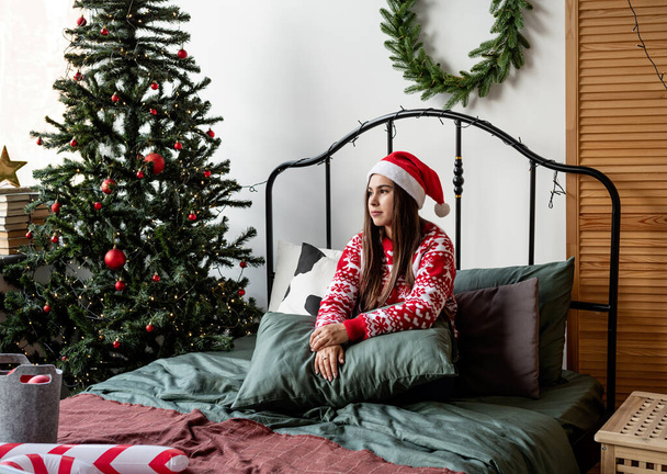 З Різдвом і Новим Роком. Щаслива молода жінка в червоному светрі і капелюсі Санти сидить на ліжку в пам'ять про хрещення - Фото, зображення