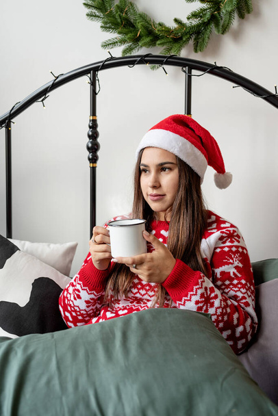 Καλά Χριστούγεννα και Καλή Χρονιά. Νεαρή μελαχρινή γυναίκα με κόκκινο πουλόβερ και καπέλο santa κάθεται στο κρεβάτι γιορτάζει τα Χριστούγεννα έχοντας φλιτζάνι ζεστό ποτό - Φωτογραφία, εικόνα