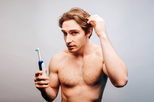 Ο άνθρωπος κρατάει την ηλεκτρική οδοντόβουρτσα και αγγίζει τα μαλλιά του - Φωτογραφία, εικόνα