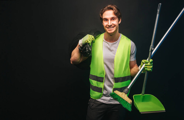 Limpiador sonriente sostiene bolsa de basura, escoba y cuchara - Foto, imagen
