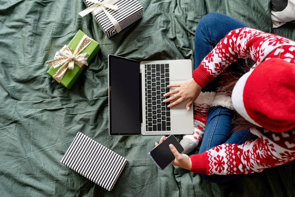 Vrolijk kerstfeest en gelukkig nieuwjaar. Online winkelen. Top view vrouw in kerstkleding zittend op het bed met behulp van laptop. Mockup scherm, kopieer ruimte - Foto, afbeelding
