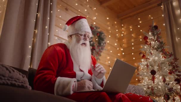 Camera beneden De Kerstman zit thuis op de bank met een laptop naast de kerstboom, werkend met een laptop - Video