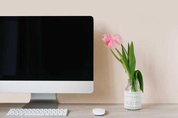 Monitor komputerowy, bukiet różowych tulipanów i notatnik z długopisem na jasnodrewnianym stole przy beżowej ścianie. Miejsce pracy wolnego strzelca lub studenta - Zdjęcie, obraz