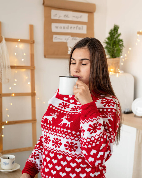 Καλά Χριστούγεννα και Καλή Χρονιά. Ευτυχισμένη νεαρή μελαχρινή γυναίκα σε χριστουγεννιάτικο πουλόβερ κάνοντας ζεστά ροφήματα στην κουζίνα - Φωτογραφία, εικόνα