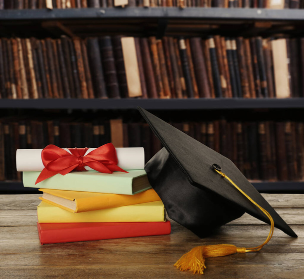 Graduation hat, books and diploma on wooden table in library - Foto, imagen