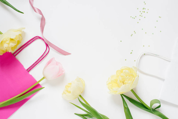 Piękna wiosna różowe i żółte tulipany kwiaty z białą wstążką z jasną torbą na białym. Leżeć płasko. Odbiór. Dzień Kobiet albo prezent na Walentynki. Koncepcja wakacyjnej wiosennej sprzedaży w sklepie lub zakupach online - Zdjęcie, obraz