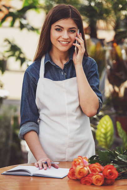 Portrait of young female florist talking on phone and making notes at flower shop. - Foto, Imagen