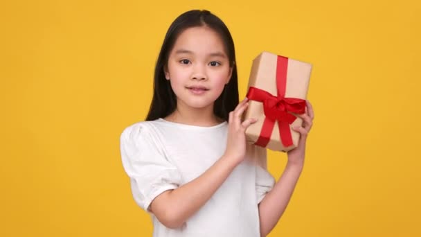 Curiosidad navideña. Adorable pequeña chica asiática sacudiendo caja de regalo, tratando de adivinar lo que está dentro, fondo de estudio naranja - Imágenes, Vídeo