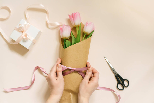 Плоскі жіночі руки зав'язують атласний стрічковий лук на простий букет свіжих рожевих квітів тюльпанів у ремісничому папері на бежевому столі
. - Фото, зображення