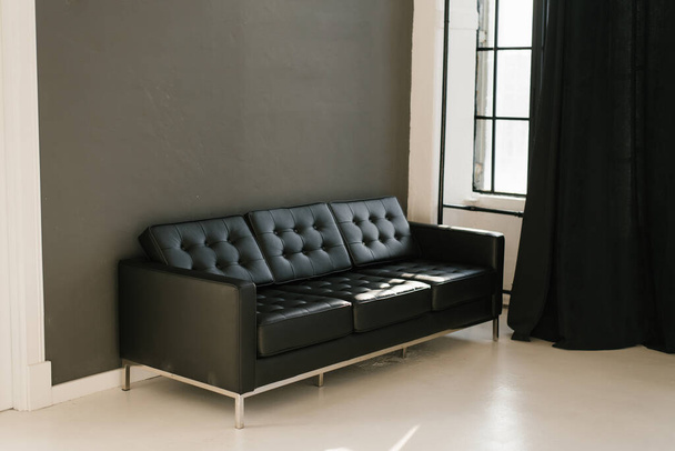Жестокий черный диван в комнате с темно-серыми стенами и черными шторами на окне - Фото, изображение
