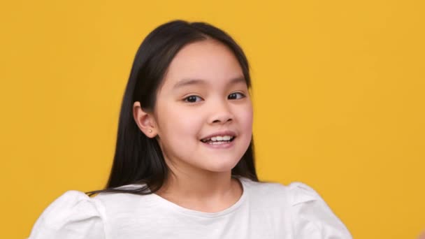 Close up portrait of adorable little asian girl showing V sign to camera and smiling widely, orange studio background - Záběry, video