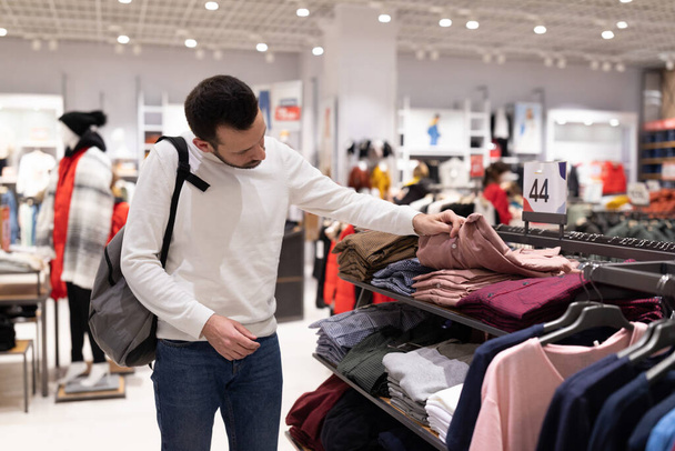 Adult handsome brunet man in white sweatshirt chooses stylish trendy shirt at clothing store in shopping mall. Shop concept - Foto, imagen