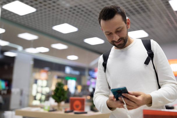 Photo of an adult positive handsome brunette man with stubble in a white sweatshirt chooses a mobile phone in an electronic store in a shopping mall - Foto, Bild