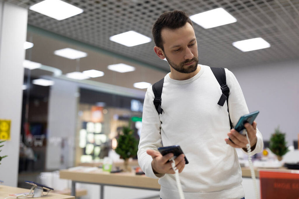 Photo of an adult doubtful handsome brunette man with stubble in a white sweatshirt chooses a mobile phone in an electronic store in a shopping mall - Photo, Image